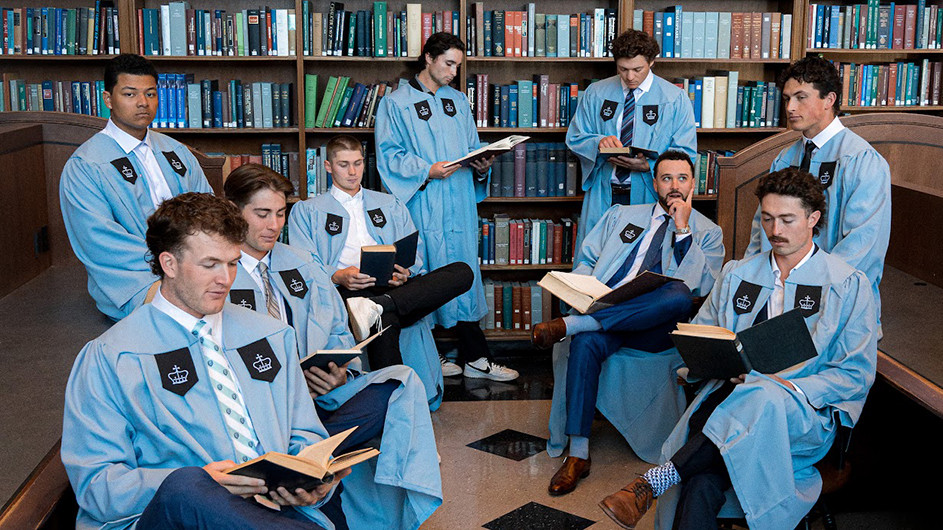 Eight men in light-blue academic-style robes with a crown-and-shield patch sit and stand in a library in front of tall bookshelves, each holding or reading a book; one sits in a chair with his hand on his chin while others look down at their books.