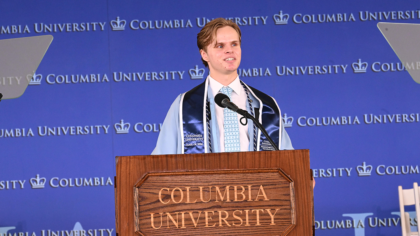 A student speaker stands at a “Columbia University” podium, wearing a stole, speaking in front of a blue Columbia University backdrop.