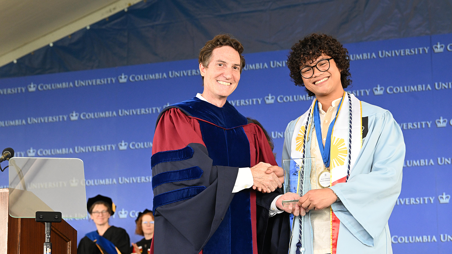 Barclay Morrison in academic regalia shakes hands with a smiling Columbia Engineering student in a light-blue cap and gown holding an award onstage in front of a Columbia University backdrop.
