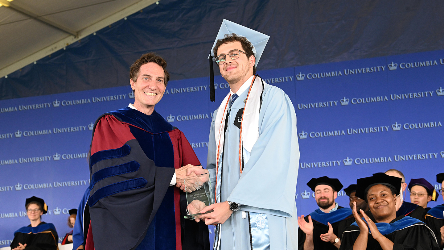 Columbia Engineering faculty member presents an award to a Columbia Engineering student in cap and gown as faculty applaud onstage.