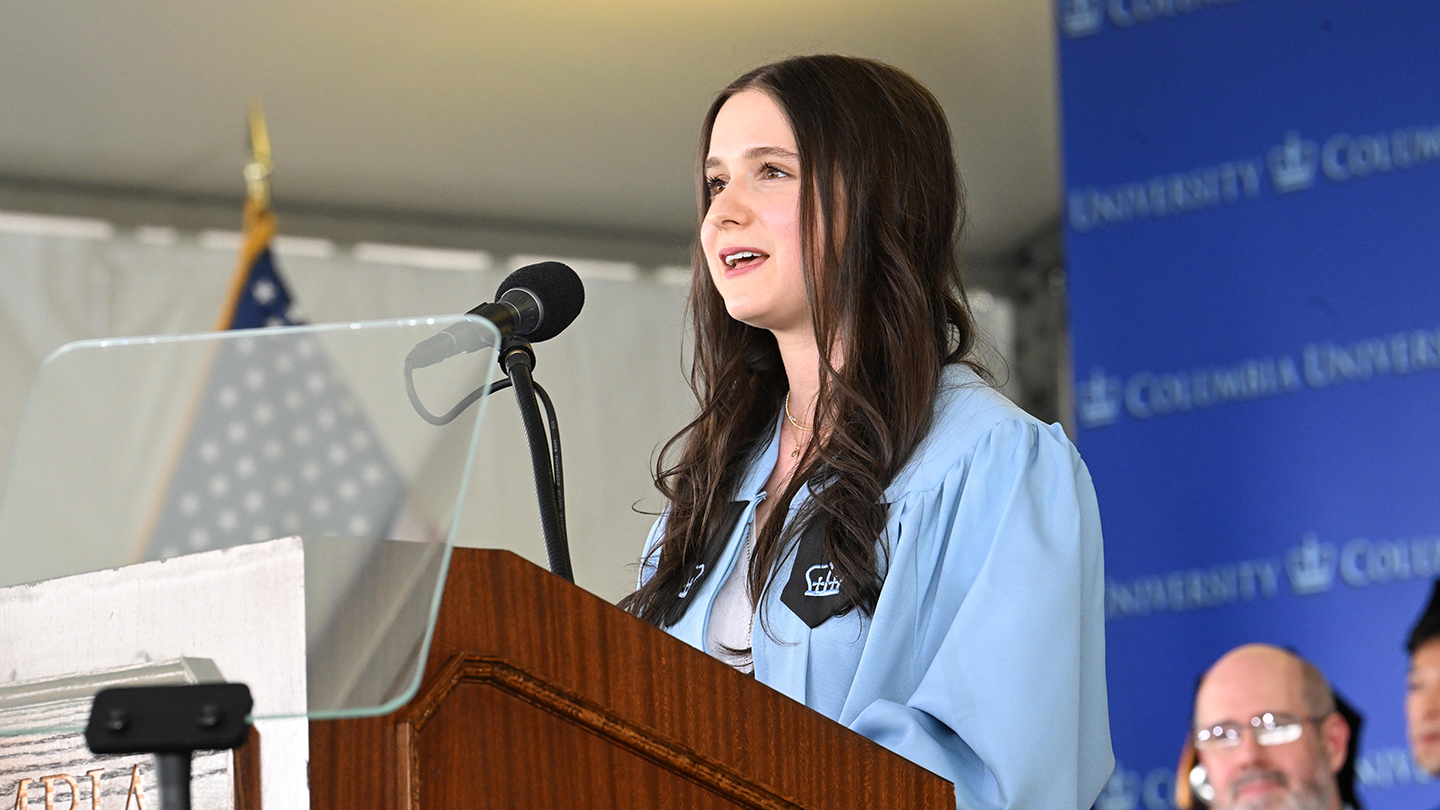 Columbia Engineering student speaker in a light-blue gown stands at a podium microphone delivering remarks during Columbia Engineering Class Day 2026.