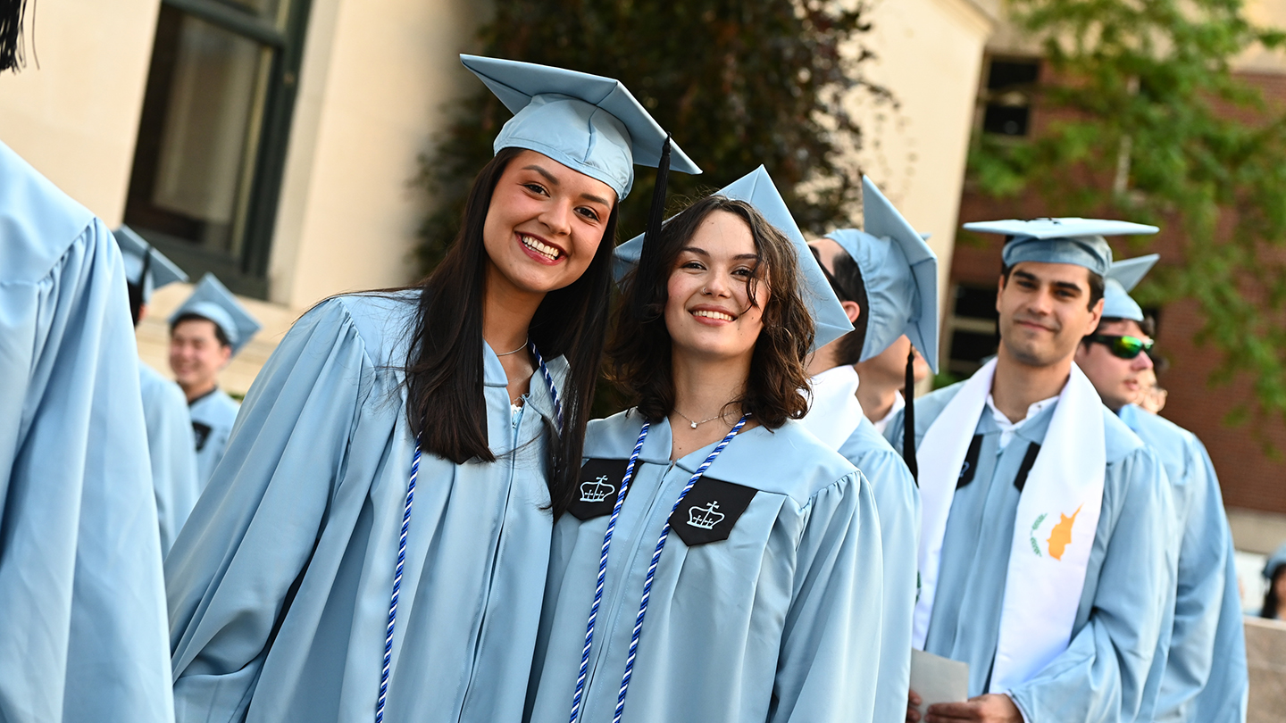 Two Columbia Engineering students in light-blue caps and gowns smile and pose together outdoors among classmates before the ceremony.