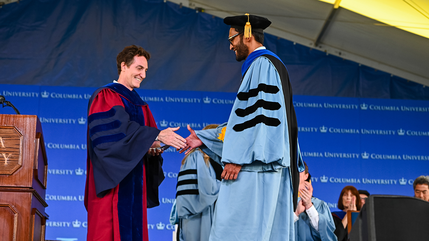 Barclay Morrison in academic regalia extends a hand to greet another faculty member onstage as the audience applauds.