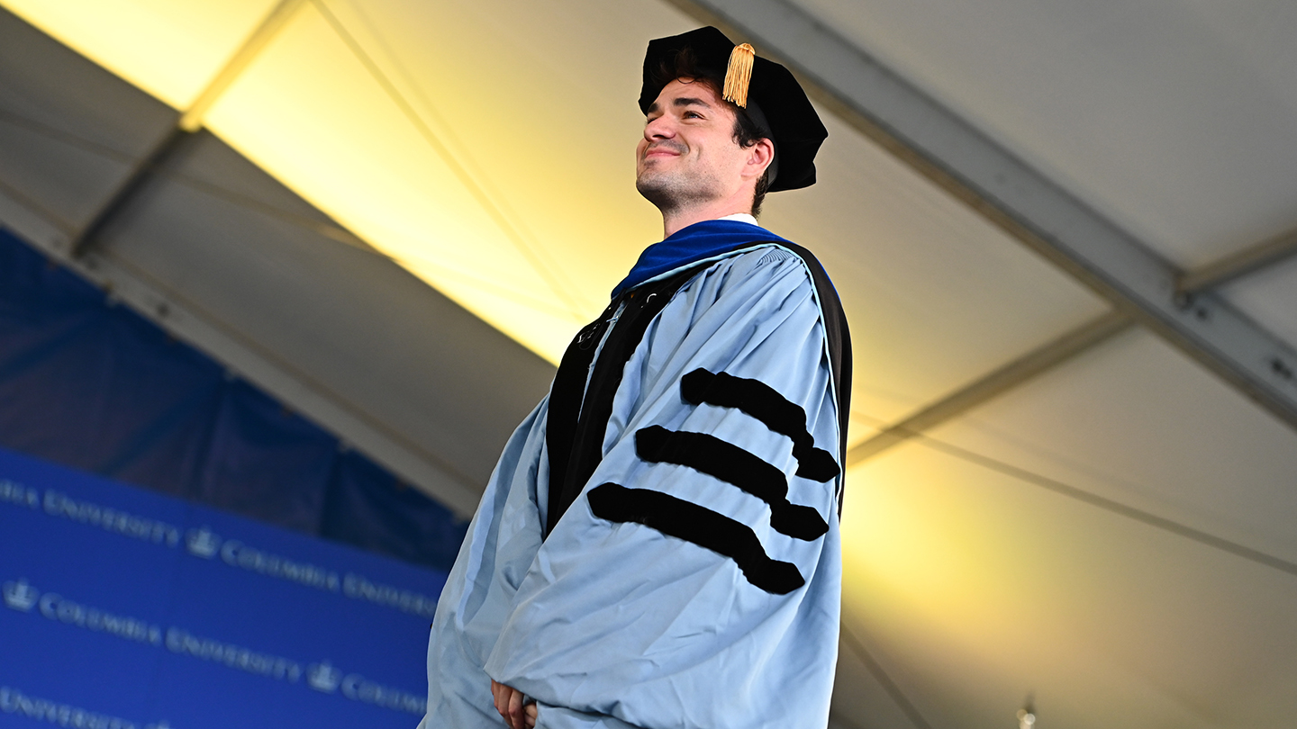 Bernard "Bert" Steyaert in light-blue doctoral regalia and tam standing onstage under the ceremony tent.