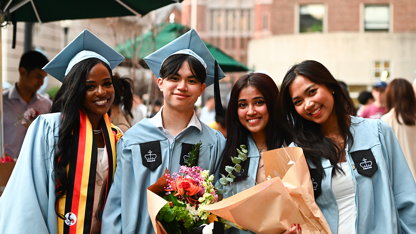 Four graduates in light-blue caps and gowns smile for a photo outdoors; two hold bouquets of flowers.