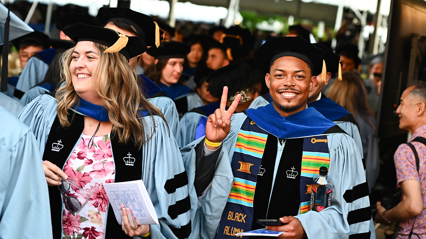 Two graduates in academic regalia stand in a crowd; one smiles and flashes a peace sign while holding a program.