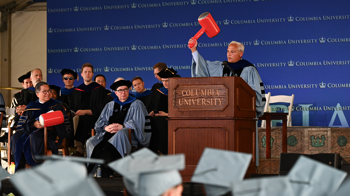 Thomas Caulfield at the podium raises a large red ceremonial mallet while Columbia Engineering students and faculty look on during the ceremony.