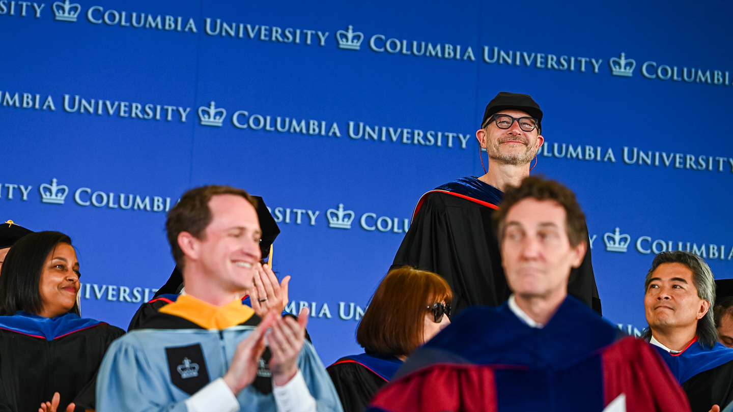 Harry West in graduation regalia standing among Columbia Engineering faculty. A blue "Columbia University" backdrop is behind him.