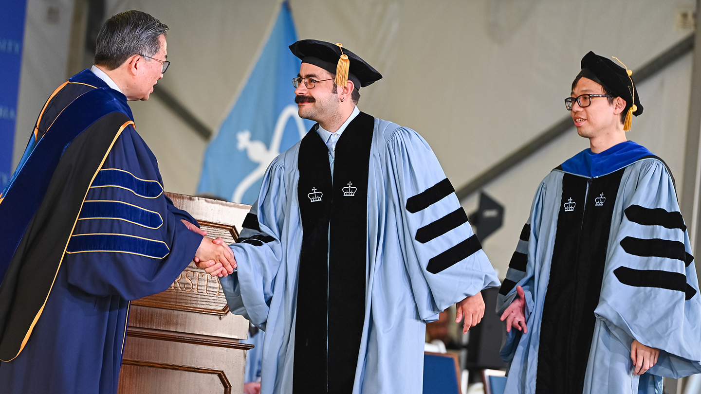 A Columbia Engineering doctoral student in graduation regalia shakes hands with Dean Shih-Fu Chang. Another doctoral student looks on from behind the other.