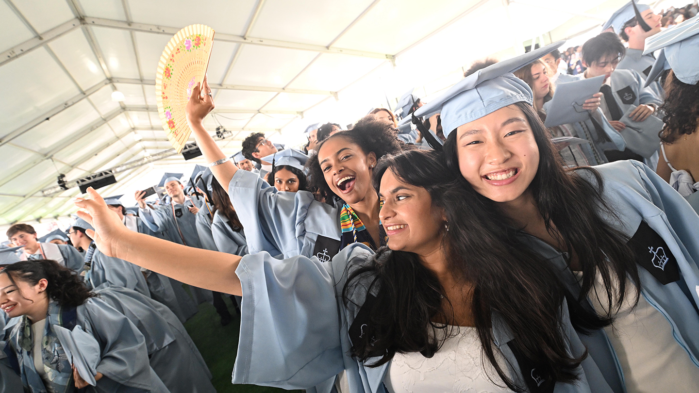 Graduates in light-blue caps and gowns take a selfie under a large tent; one person raises a decorative fan.