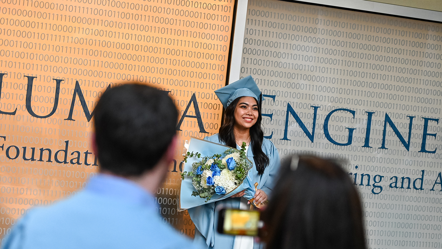 A Columbia Engineering student in graduation regalia holding a bouquet of flowers in front a sign that reads "Columbia Engineering". 