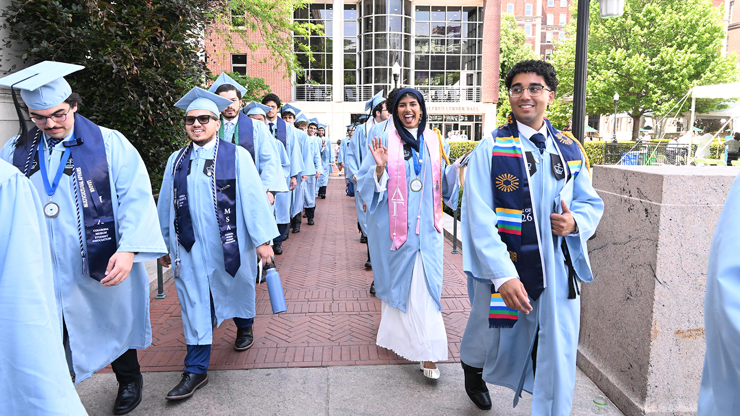 A line of graduates in light-blue caps and gowns walks along a campus path toward a ceremony area.