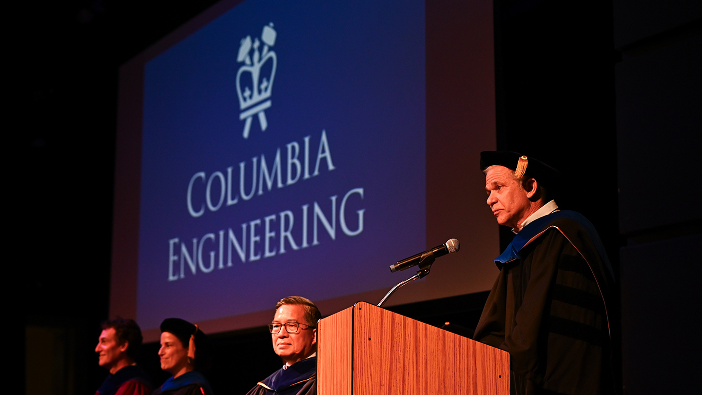 Michael Weinstein in graduation regalia speaking into a microphone on stage. The projection screen behind her reads "Columbia Engineering"