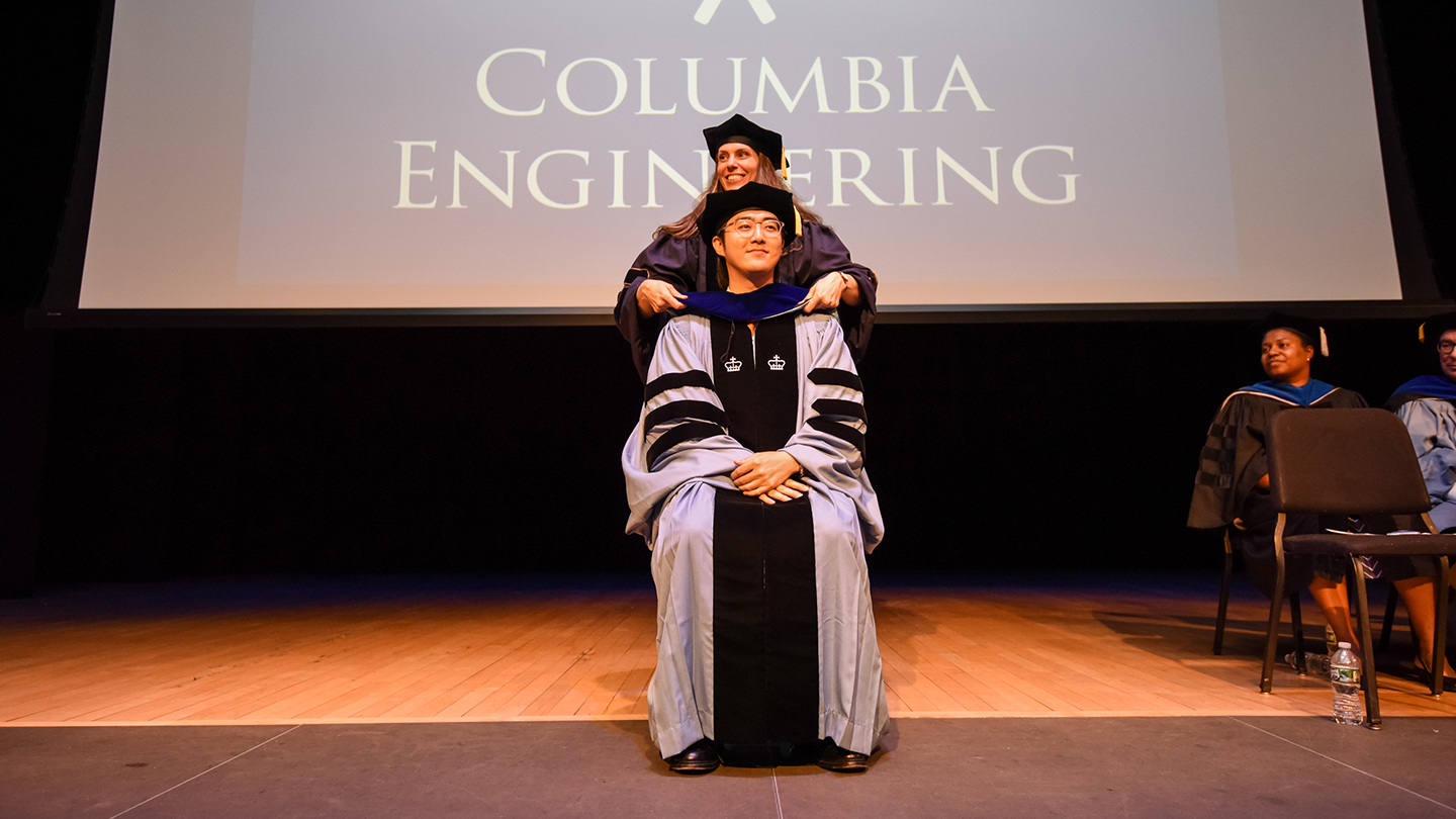 A Columbia Engineering doctoral graduate in regalia sitting in a chair. A faculty member stands behind them adjusting their hood. 