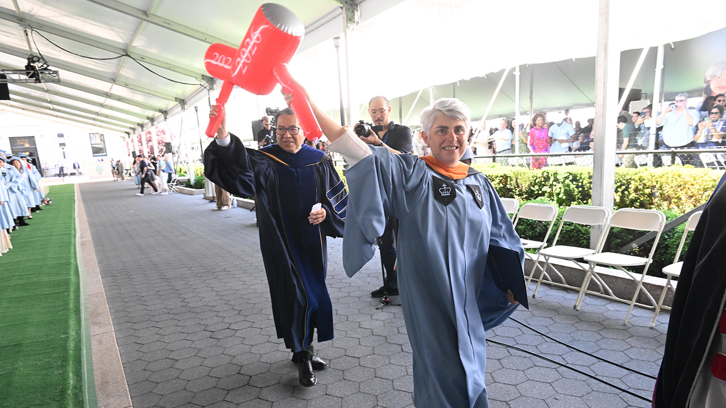 Two faculty or staff in academic robes walk under a tent, each holding an oversized red inflatable gavel marked “2026.”