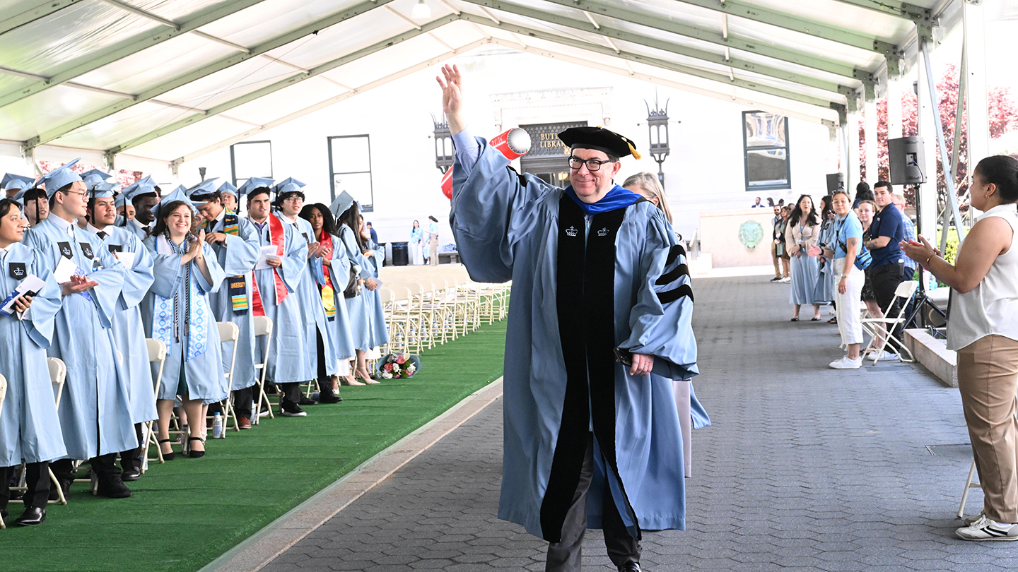 A faculty member in academic regalia waves while walking down the aisle as graduates and guests applaud.