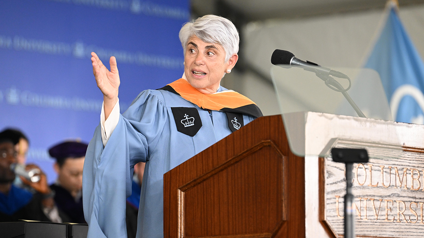 Angeliki Frangou in academic regalia gestures while addressing the audience from a podium with a “Columbia University” backdrop.