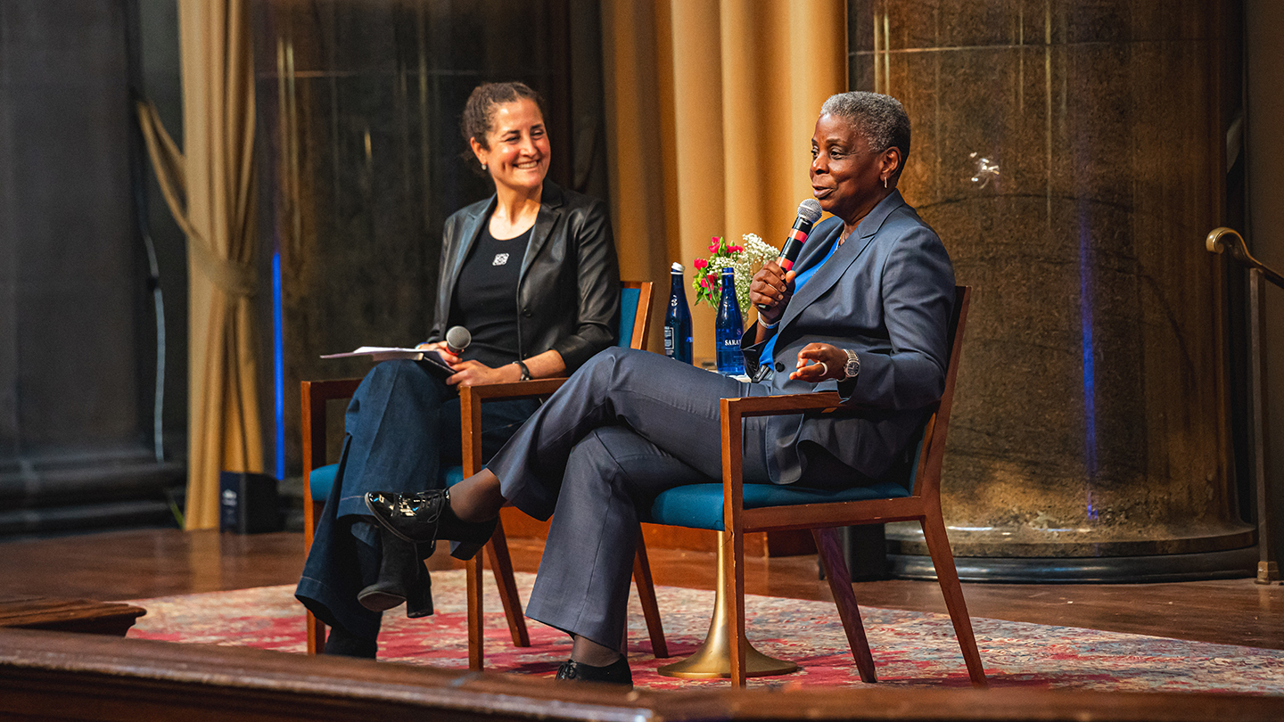 Two speakers sit onstage in armchairs during a talk; one woman speaks into a handheld microphone while the other smiles and listens.