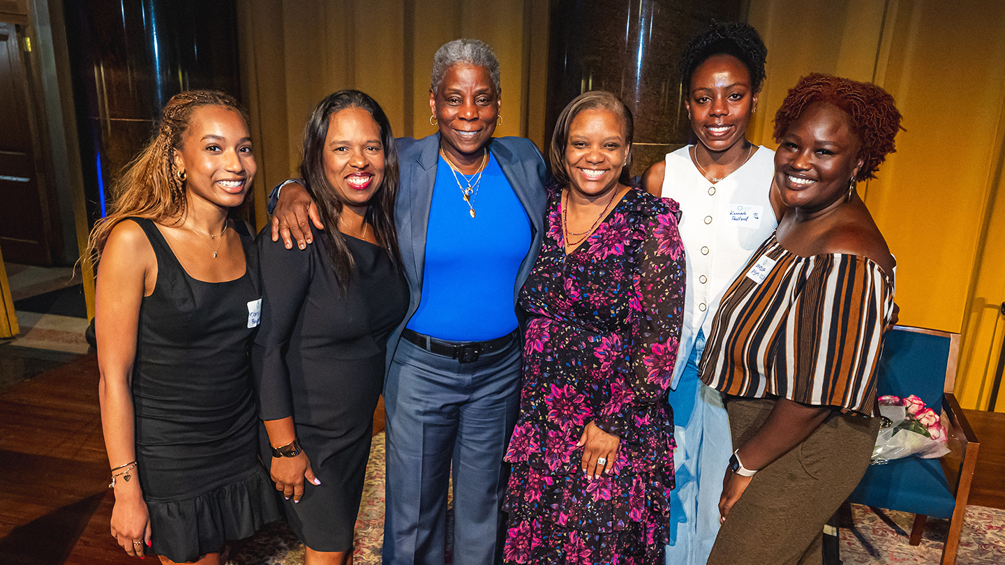 Six attendees stand arm-in-arm and smile for a group photo at an indoor event venue.