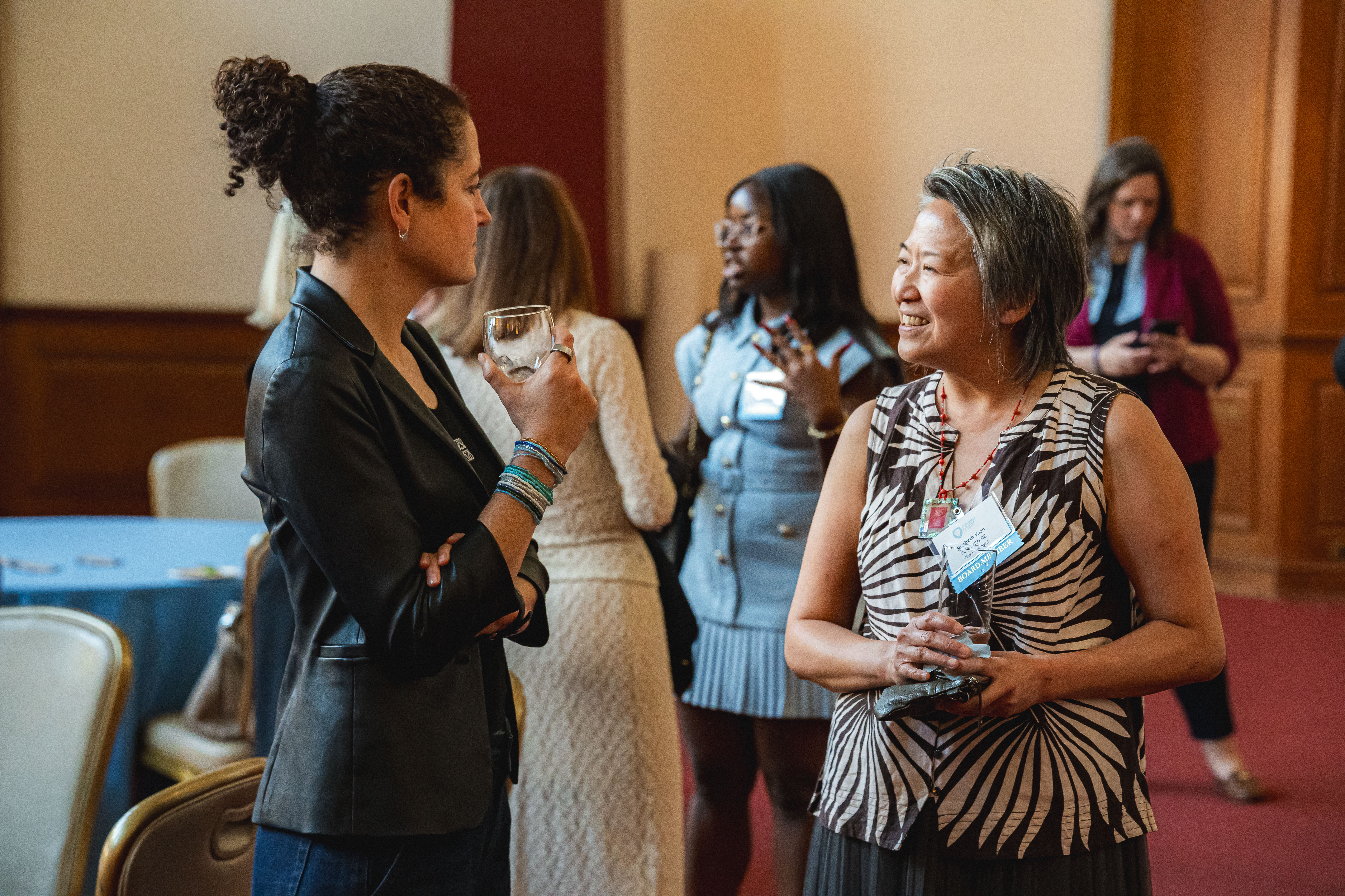 Two attendees talk during a reception; one holds a drink while the other smiles, with other guests mingling in the background.