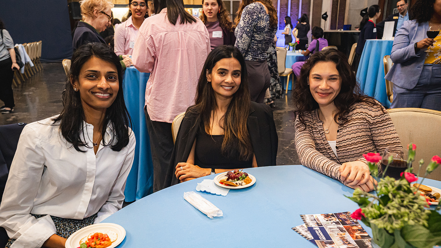 Three attendees sit at a round table during a reception, smiling toward the camera with small plates of food on the table.