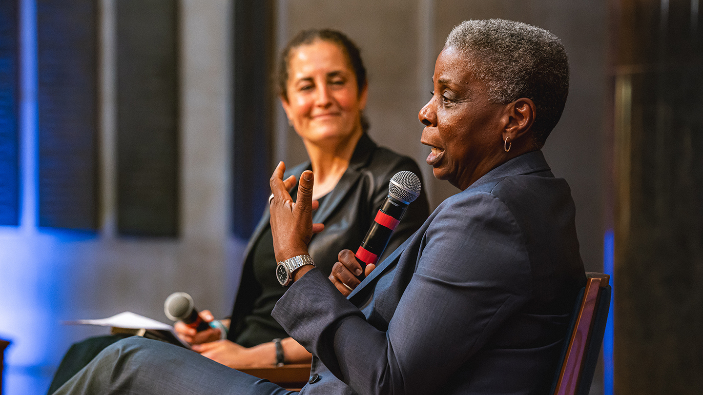 Close-up of a speaker holding a microphone and gesturing while another seated moderator listens in the background.