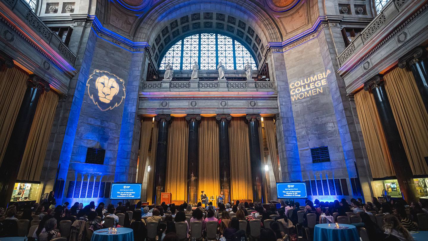 Wide shot of a large hall with a stage, audience seating, and “Columbia College Women” projected on the walls.