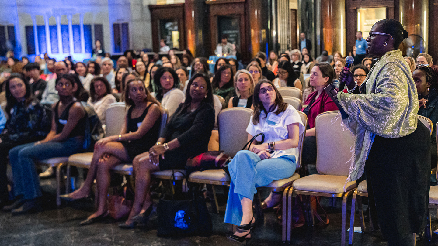 A woman stands in the aisle speaking into a microphone as seated attendees listen.