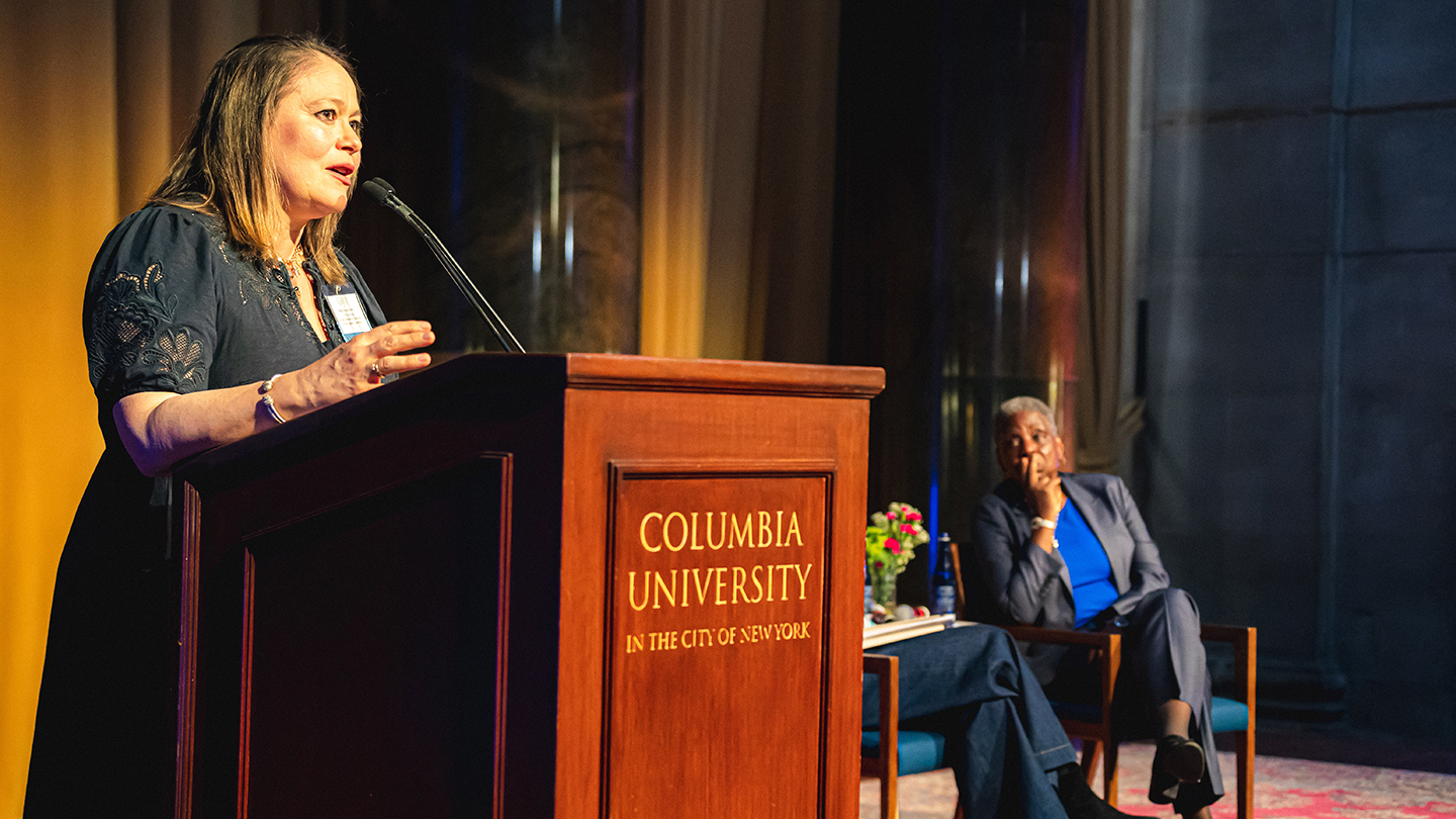 A woman speaks at a podium labeled “Columbia University” while another speaker sits onstage behind her.