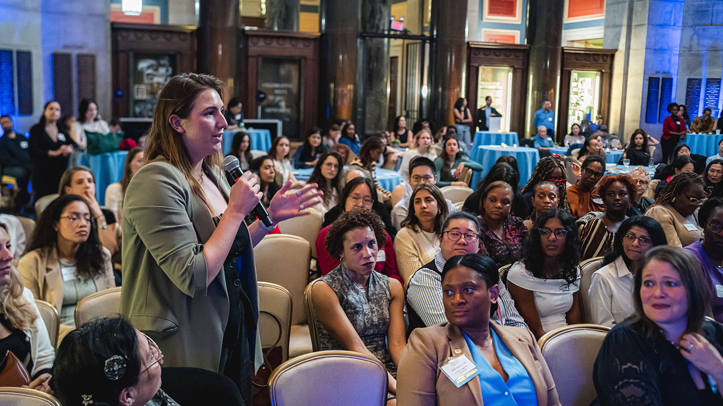 An audience member stands and asks a question into a microphone in a crowded auditorium.