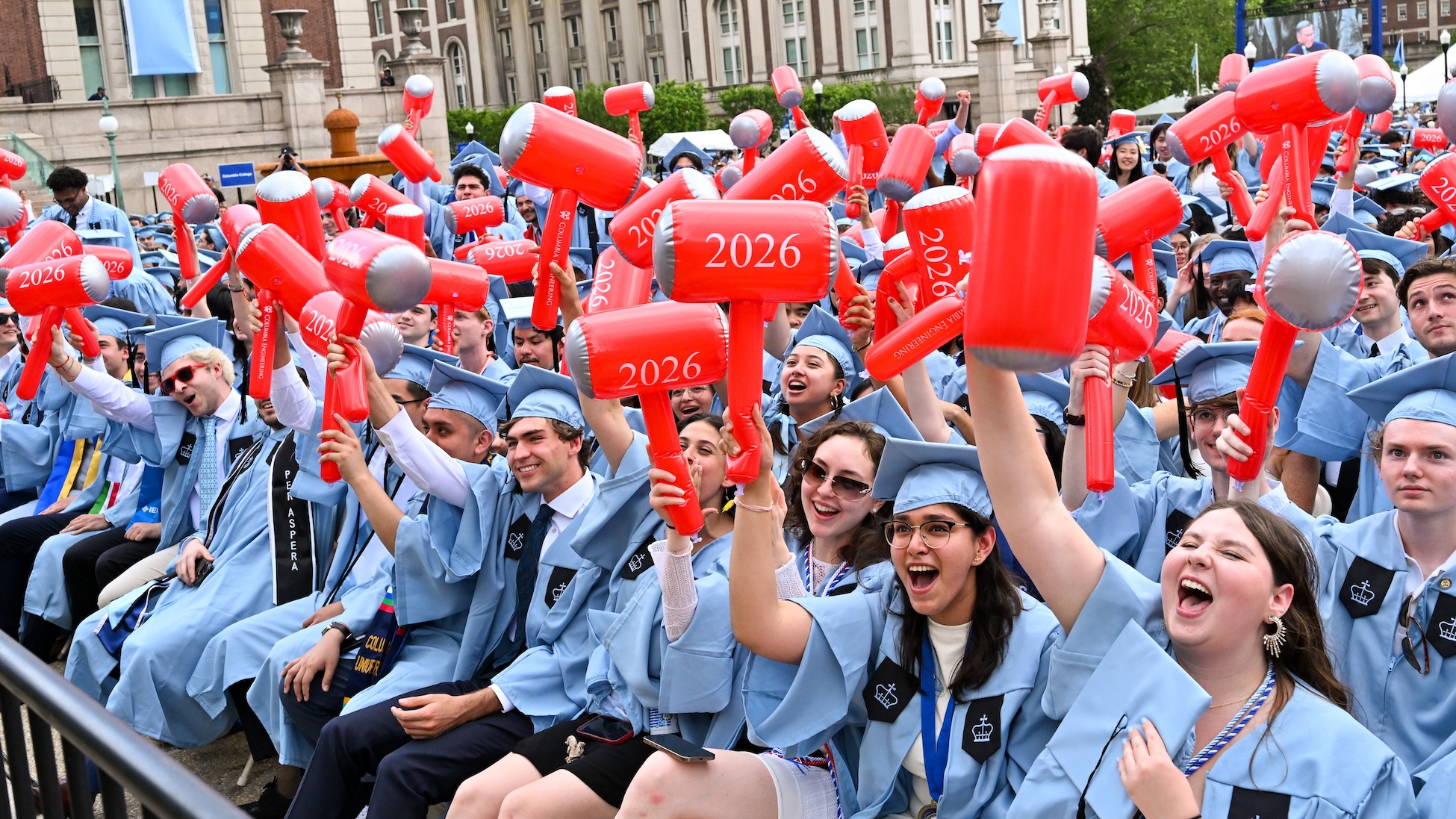 A large crowd of Columbia Engineering graduates in light blue caps and gowns cheer and wave red inflatable mallets printed with “2026” during an outdoor graduation ceremony