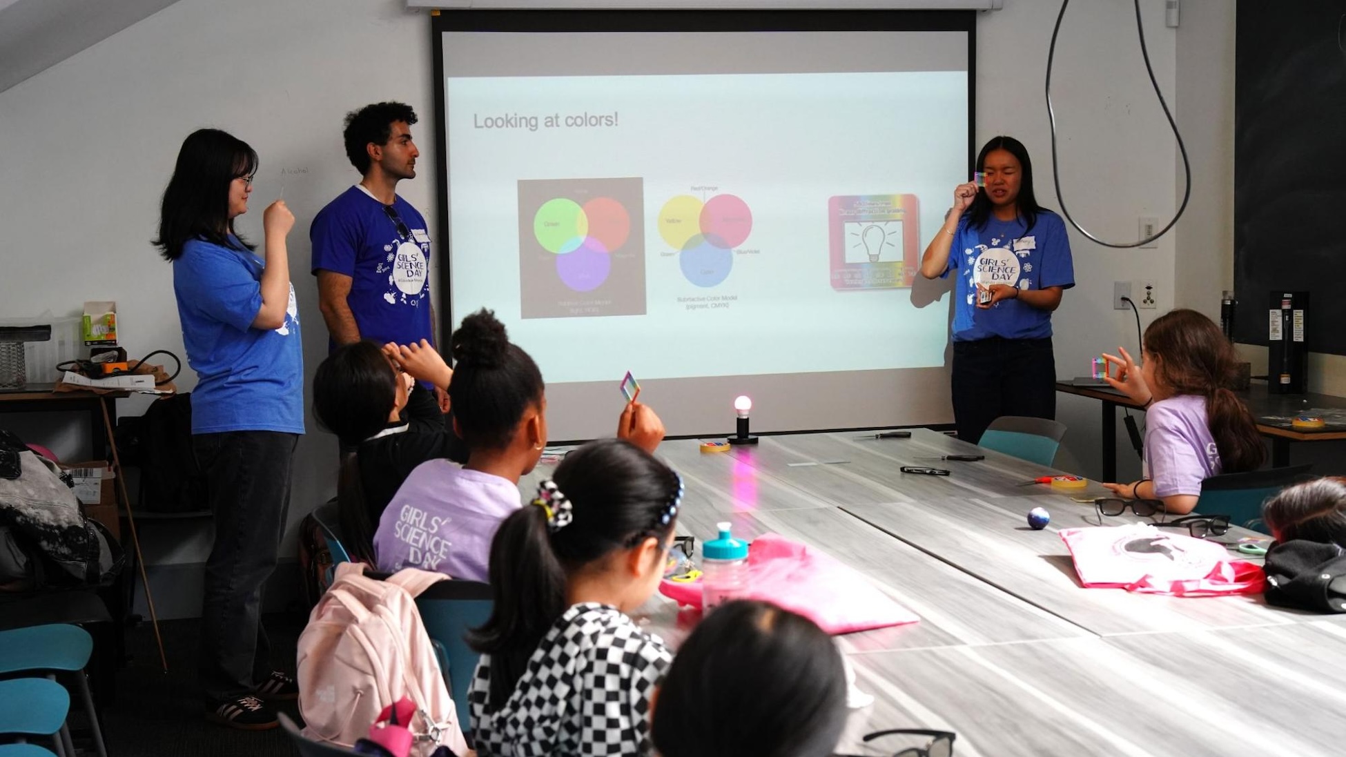 In a classroom, three instructors in blue “Girls Science Day” T-shirts lead a lesson on color. A projector screen shows a slide titled “Looking at colors!” with overlapping color-circle diagrams, while several students sit around a long table holding small prisms or color filters and watching a glowing light in the middle of the table.