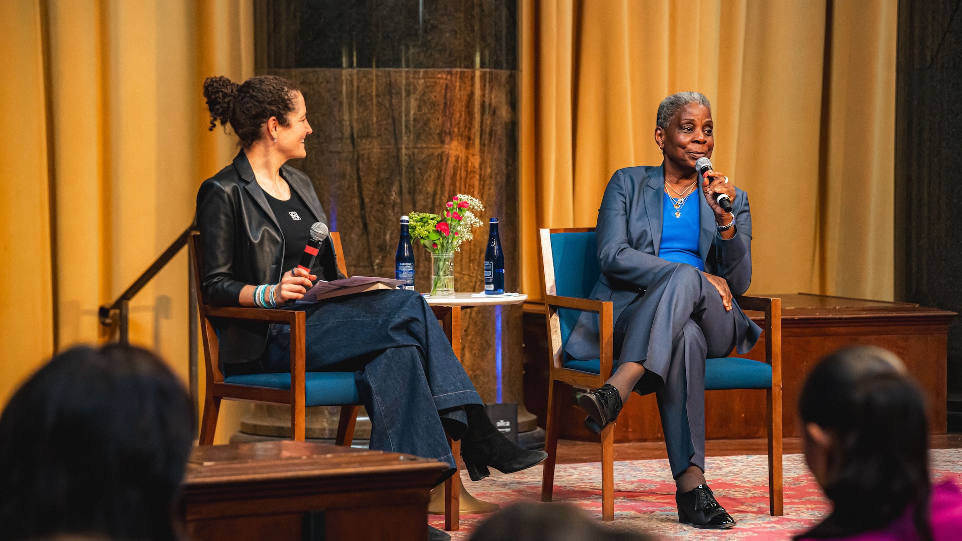 Two women sit on stage in armchairs during a public talk; one listens with a microphone in hand while the other, in a blue suit, speaks into a microphone. A small table between them holds water bottles and a vase of flowers, with an audience visible in the foreground.
