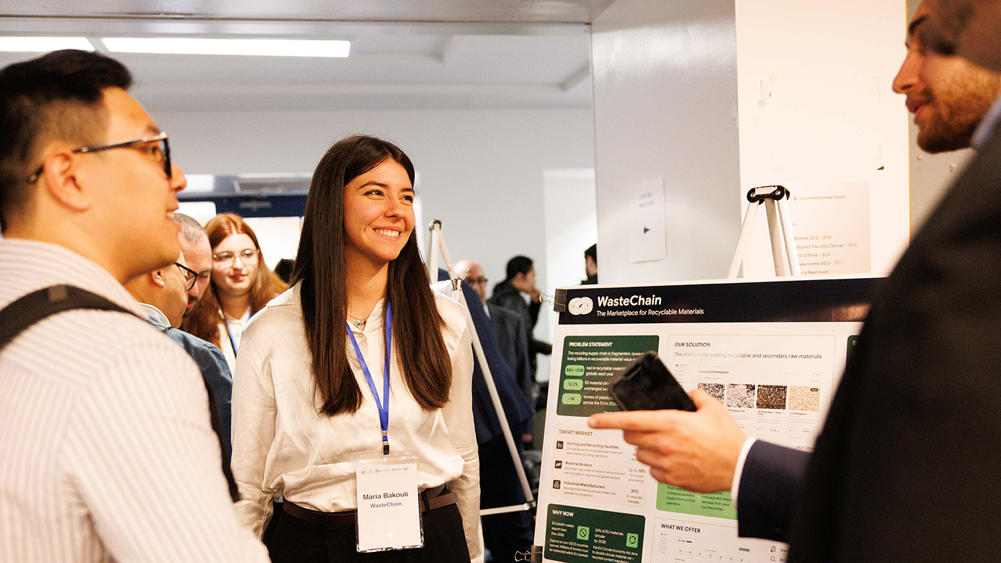 A conference attendee smiles while discussing a “WasteChain” poster with other attendees in a hallway poster session.