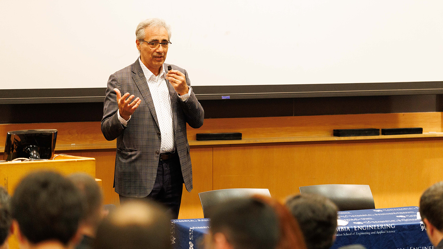 A speaker stands in front of a projection screen, holding a handheld microphone while presenting to an audience.
