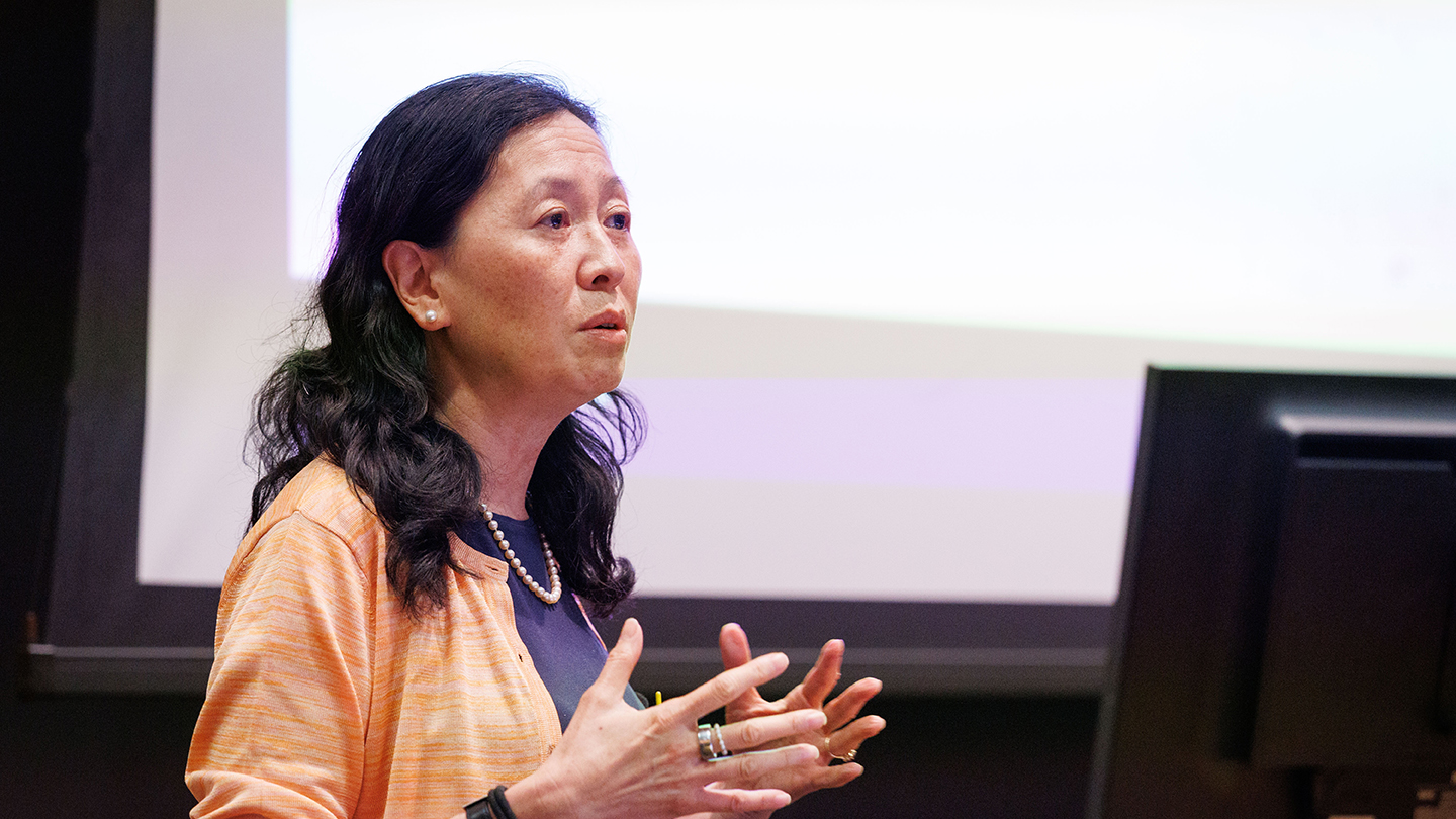A presenter gestures with both hands while speaking beside a large projection screen.