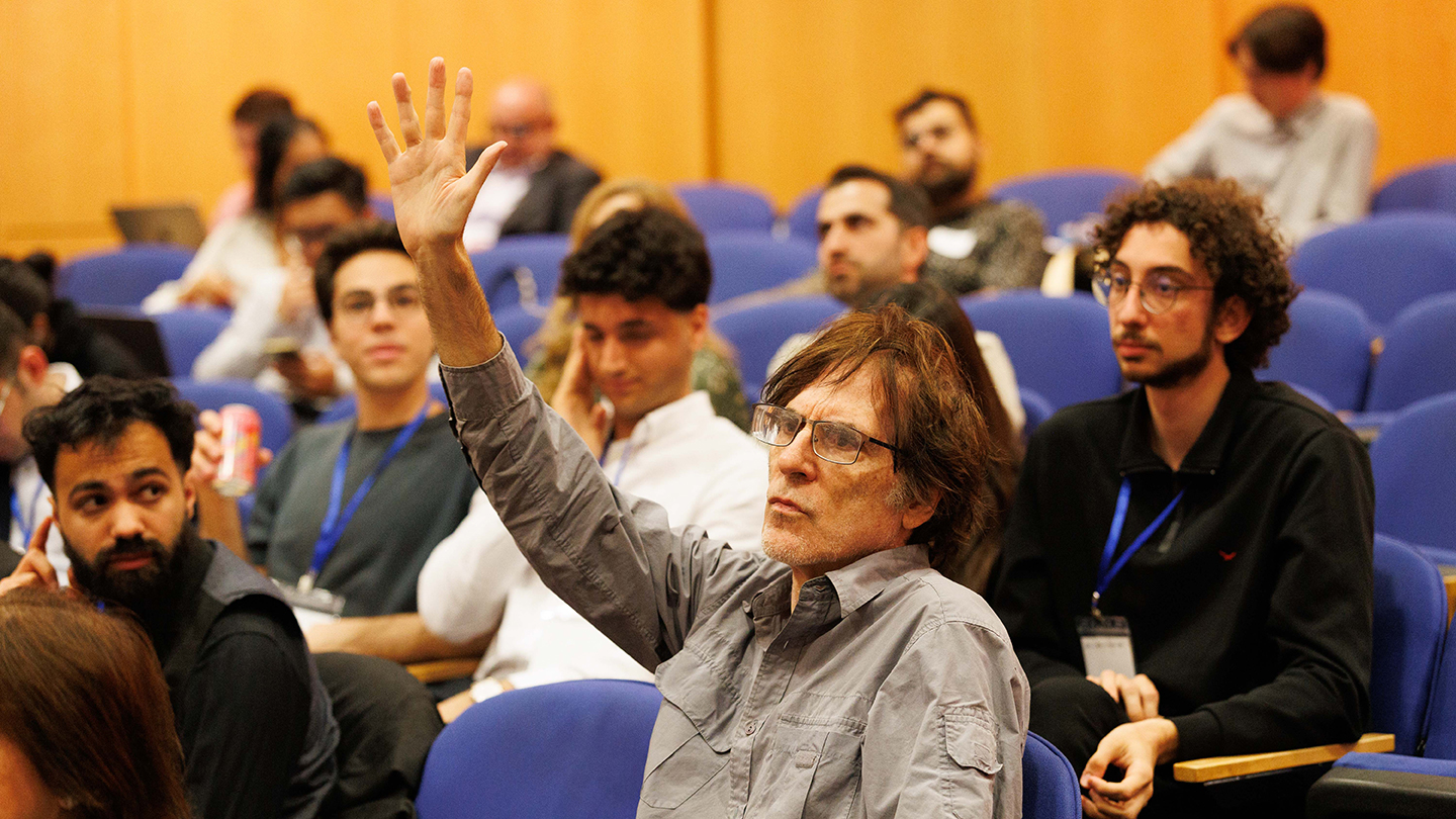 An audience member raises his hand to ask a question in a lecture hall.