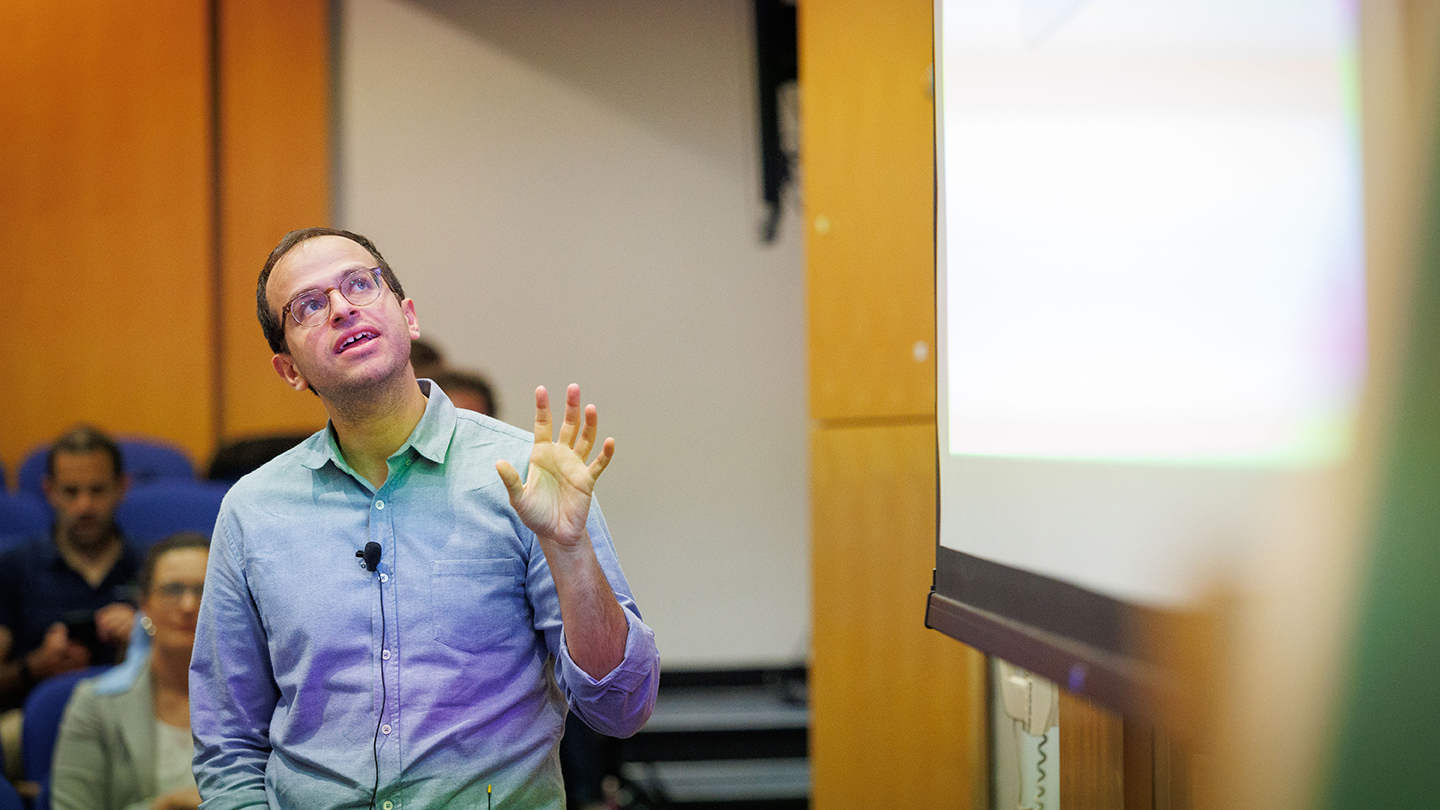 A speaker in a button-down shirt gestures while looking up at a projected slide during a talk.
