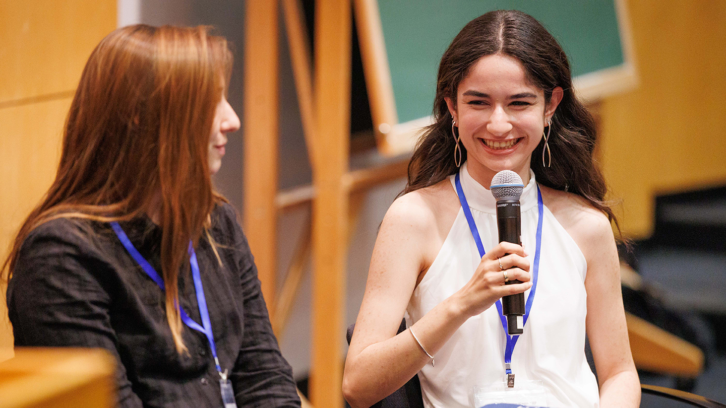 A panelist in a white sleeveless top smiles while holding a microphone during a discussion.