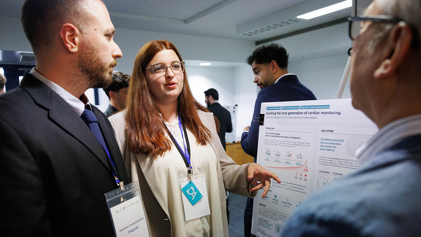 At a poster session, an attendee points to a research poster about cardiac monitoring while speaking with two other attendees.