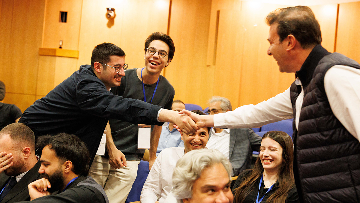 An attendee leans across seats to shake hands with another attendee as people around them smile.