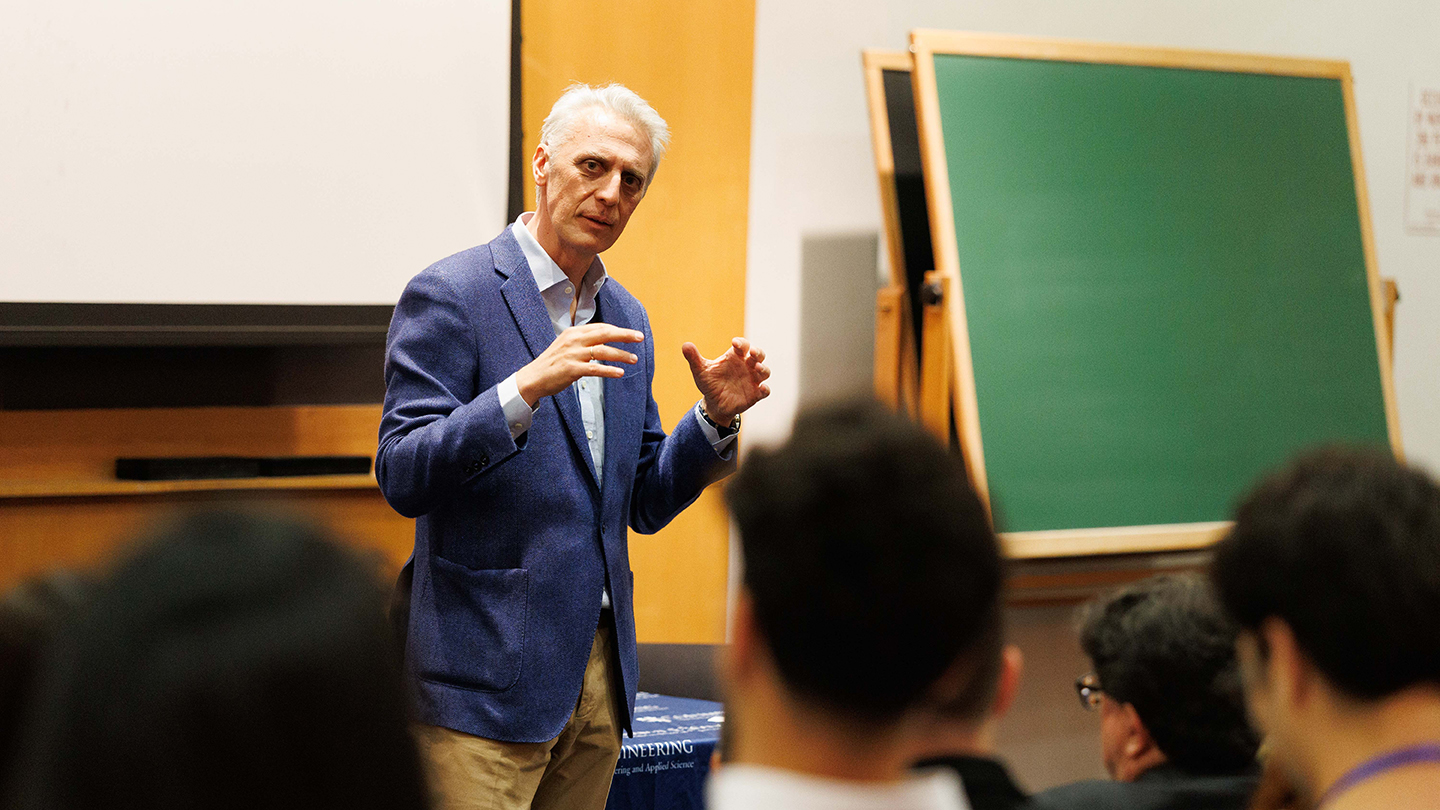 A speaker in a blue blazer gestures while presenting to an audience; a chalkboard stands behind him.