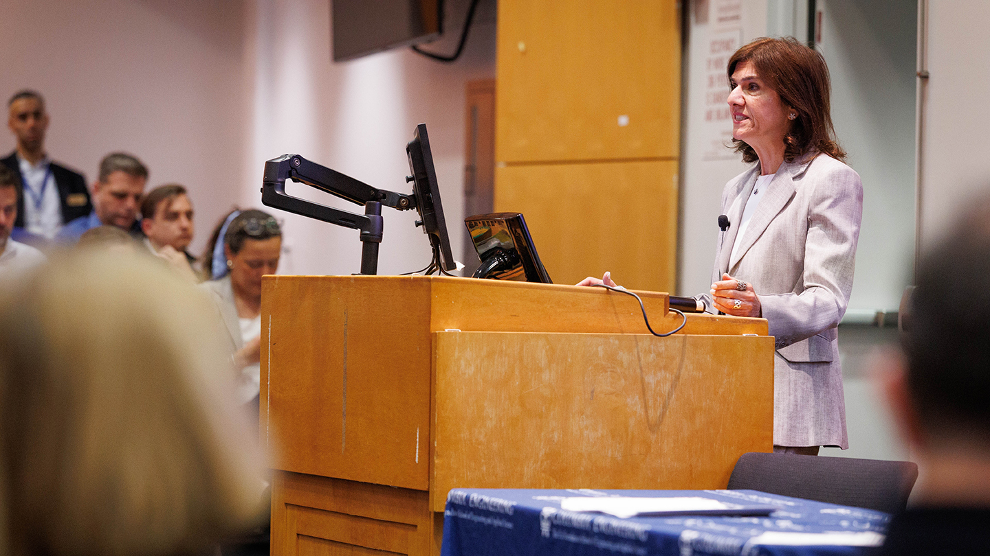A woman speaks at a podium with a computer monitor, addressing an audience seated in a lecture hall.