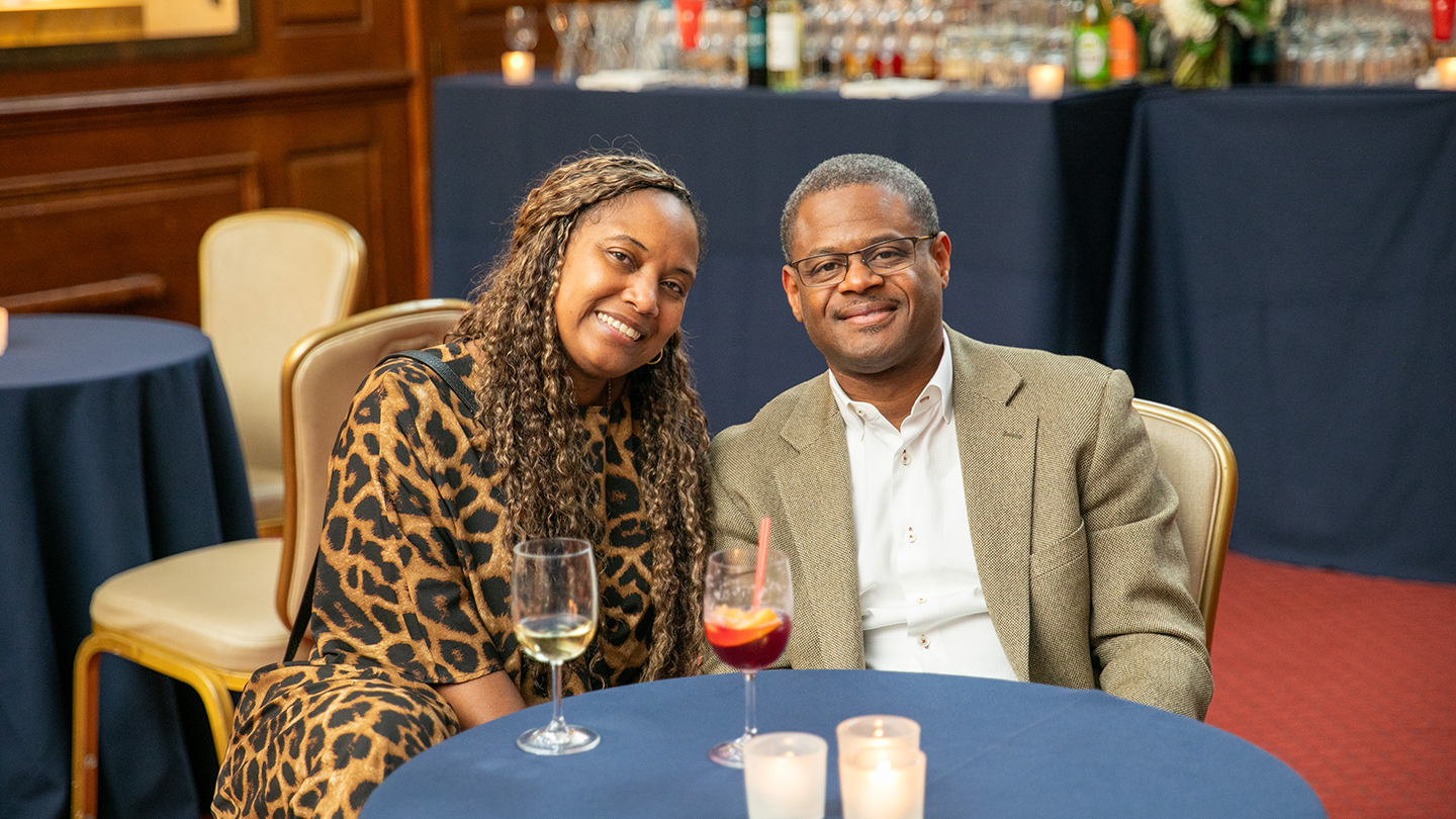 Two Columbia Engineering alumni sit close together at a cocktail table, smiling at the camera, with wine glasses and candles on the table and a bar set up behind them.