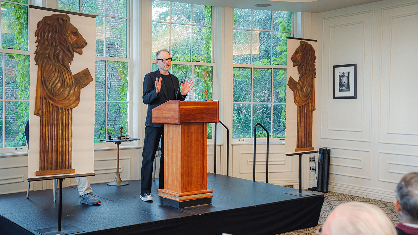 Harry West stands behind a podium in a bright room with large windows, speaking between two tall banners featuring carved lion figures during a Columbia Engineering alumni reunion program.