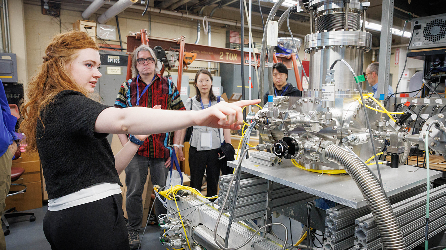 A guide points to a large piece of scientific equipment during a lab tour while several alumni watch in the background.