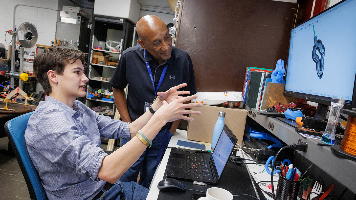In a workshop lab, a seated student gestures while explaining a 3D model on a computer monitor to an alumnus standing beside them.