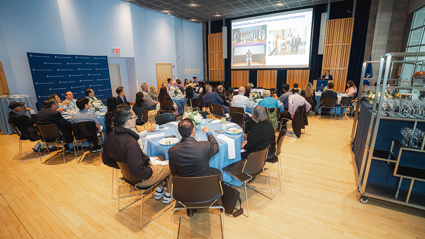 Alumni sit at round tables listening to a speaker at a podium during a reunion luncheon; a presentation slide is projected at the front of the room.