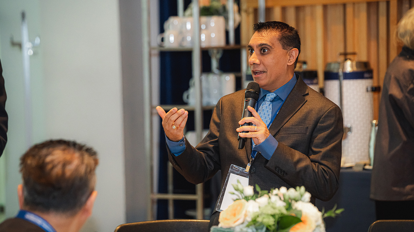  A man in a suit speaks into a handheld microphone and gestures with his free hand during an indoor reunion event.