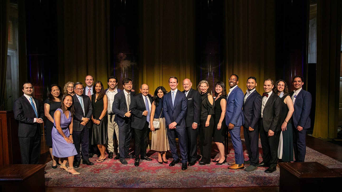 A large group of people pose on a stage in formal attire in front of tall gold curtains.